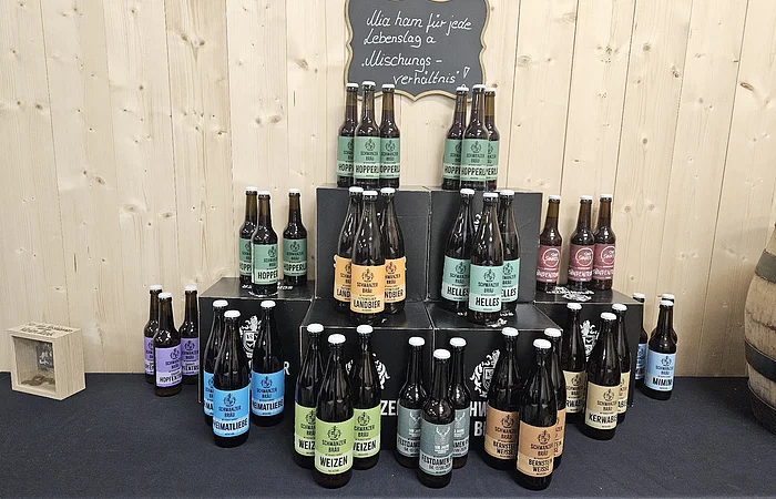 Mehrere Bierflaschen verschiedener Sorten stehen gestapelt auf schwarzen Kisten vor einer Holzwand mit einem kleinen Schild darüber.