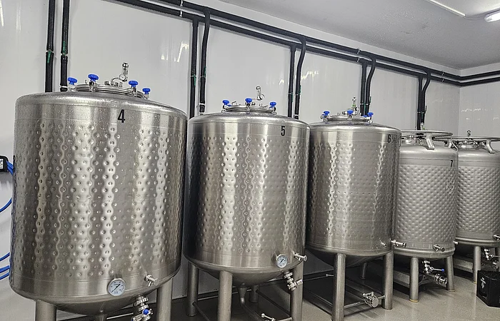 Fünf nummerierte silberne Edelstahlfässer mit Ventilen und Schläuchen in einem weißen Raum nebeneinander.