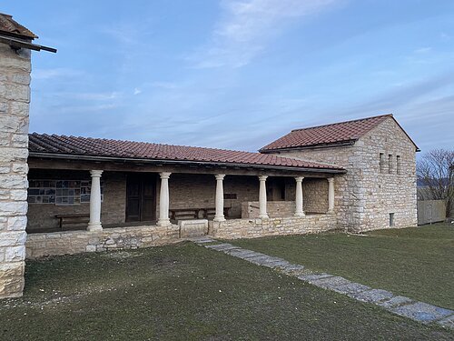 Steinernes Gebäude mit Säulen und rotem Ziegeldach vor bewölktem Himmel und Rasenfläche