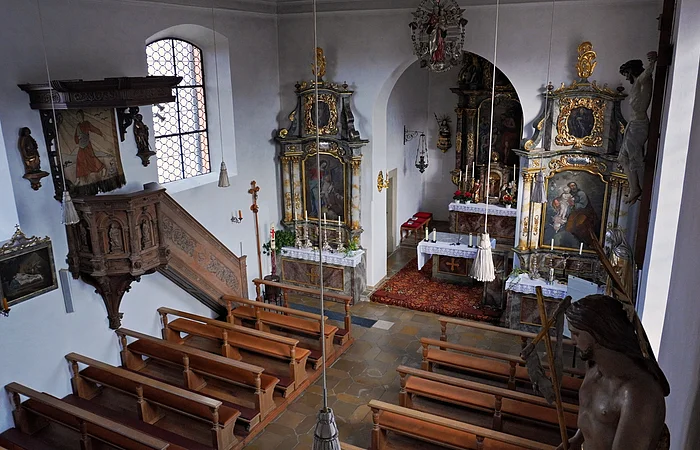 Innenraum einer kleinen Kirche mit Holzbänken, Kanzel, Altären und religiösen Gemälden an den Wänden.
