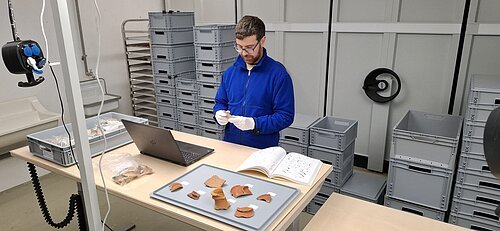 Mann mit blauer Jacke untersucht Keramikfragmente an einem Tisch mit Laptop und Buch in Lagerraum.